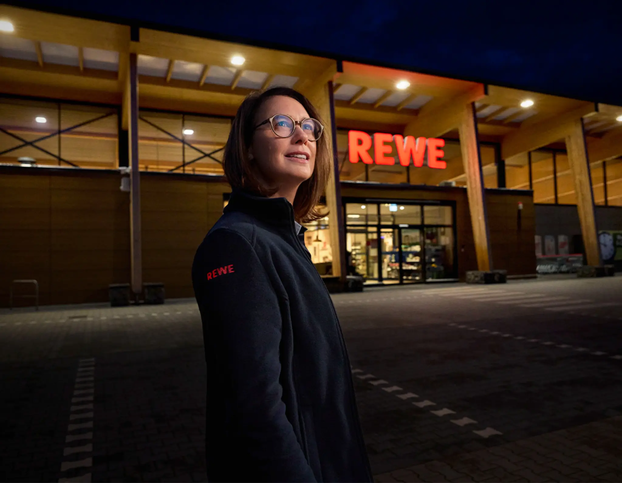 Ein beleuchteter REWE Markt am Abend. Davor steht eine Person, die bei REWE arbeitet. 
