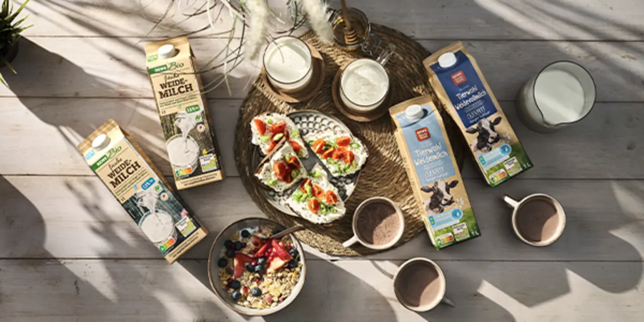 Verschiedene REWE Produkte aus höheren Haltungsstufen liegen auf einem Tisch. Dazwischen stehen Gläser voll Milch und eine Müslischüssel. 