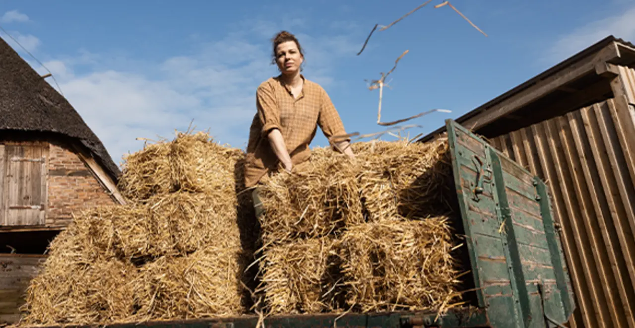 Ein Landwirtin steht oben auf einem Heuwagen und hebt einen Ballen Heu an um ihn abzuladen.  