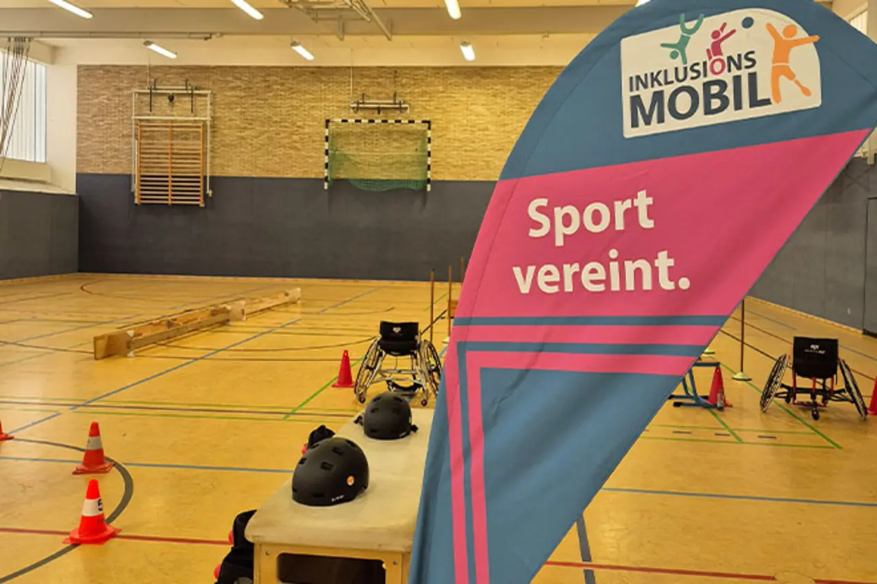 Sporthalle mit aufgebautem Rollstuhl-Parcours, Hütchen und Helmen auf einem Tisch; im Vordergrund ein Banner mit der Aufschrift ‚Inklusionsmobil – Sport vereint. 