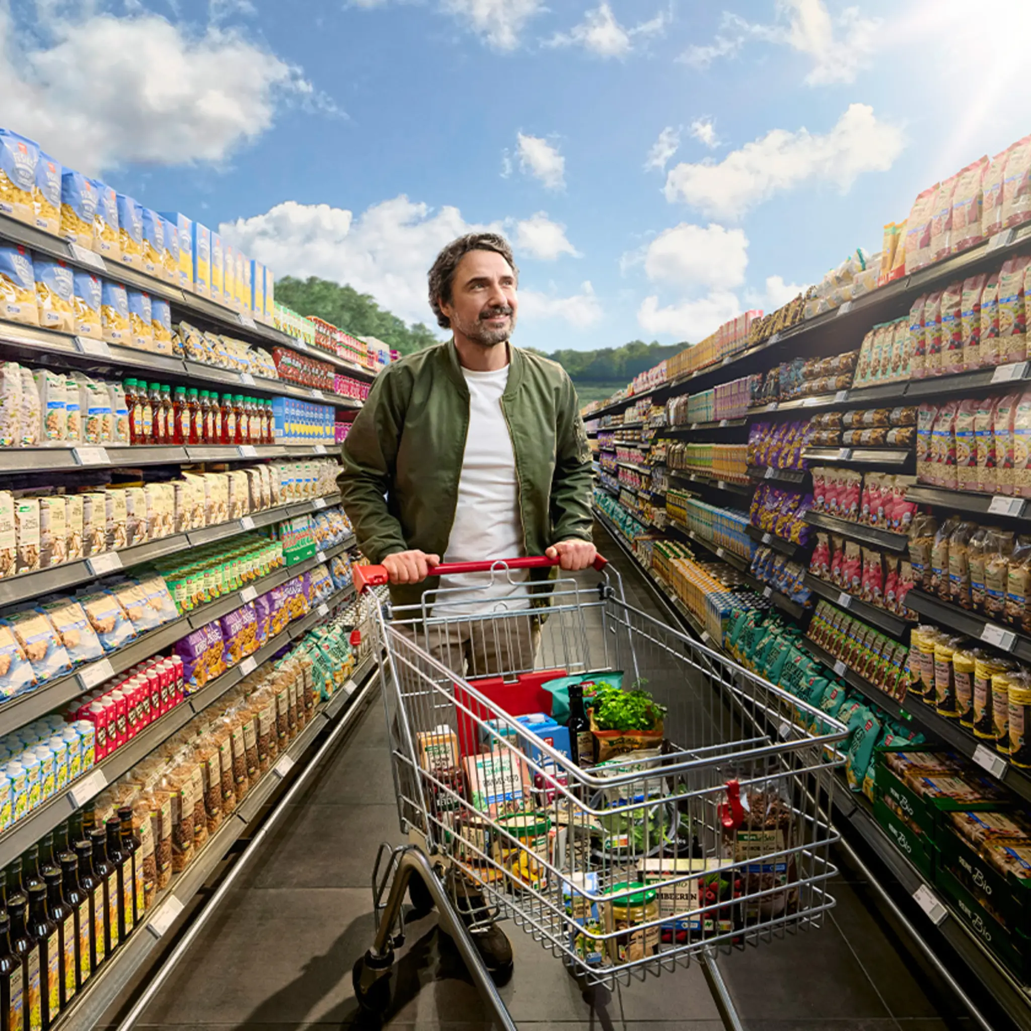 Hinter einer scheinbar endlosen Regalreihe beginnt eine Wiese mit einem Waldstück. Ein REWE Kunde betrachtet die riesige Auswahl an nachhaltigeren Produkten. 