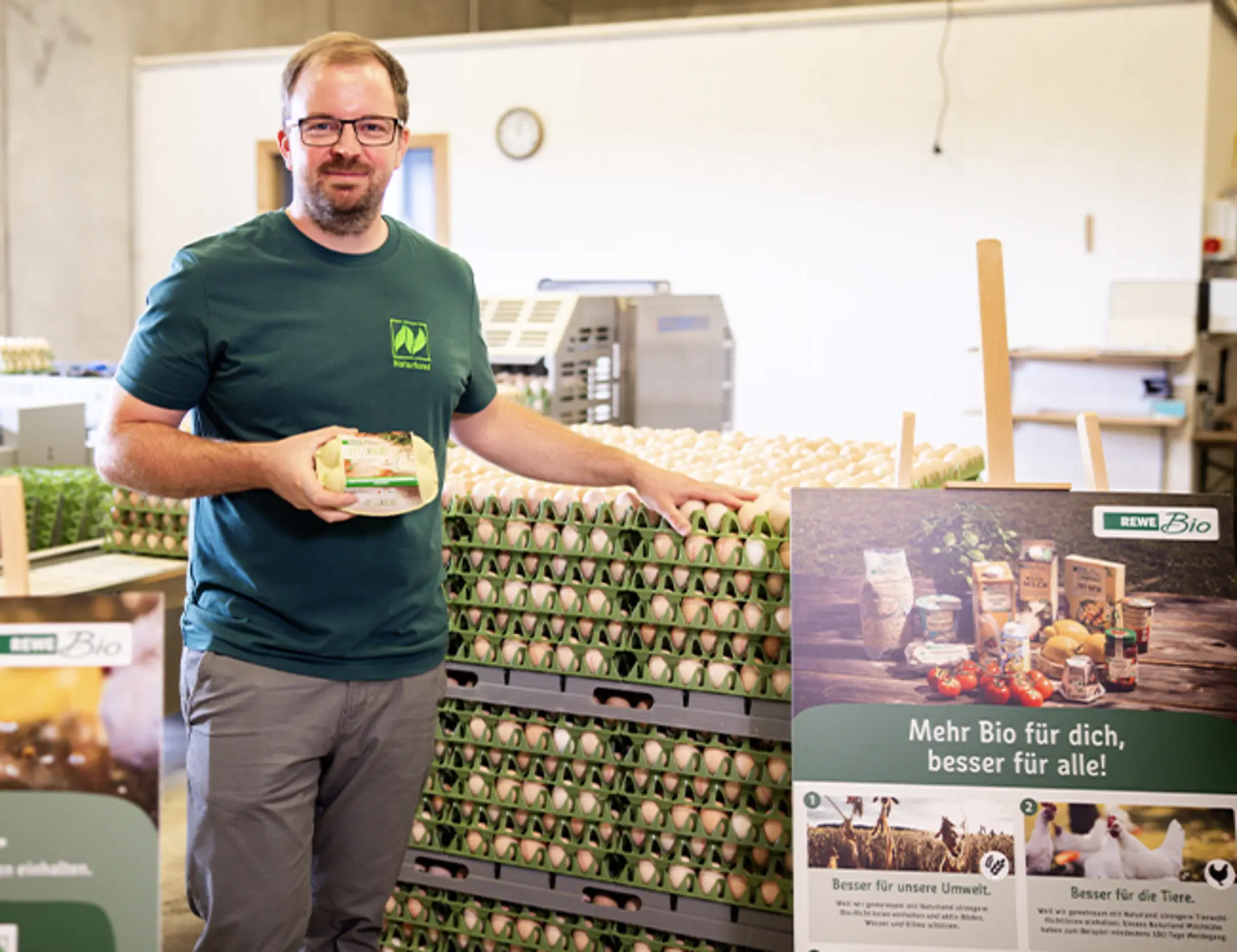 Christoph Möller hält eine Packung REWE Spitz & Bube Eier in der Hand. Ein großes Plakat von REWE Bio trägt die Aufschrift „Besser für unsere Tiere“.