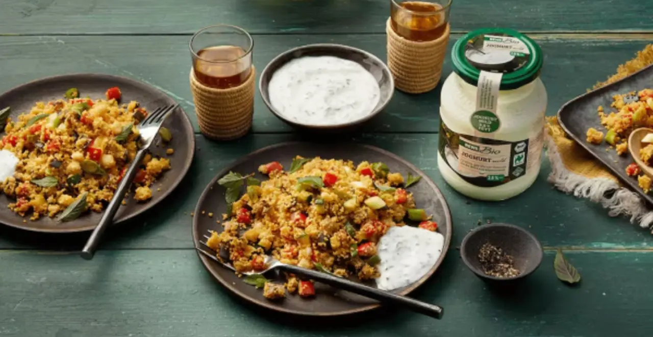 Ein Teller Couscous mit Gemüse und Joghurt-Kräuter-Soße. Oberhalb eine Platte mit Couscous und eine Schüssel mit Soße. Links daneben ein Glas REWE Bio Joghurt.