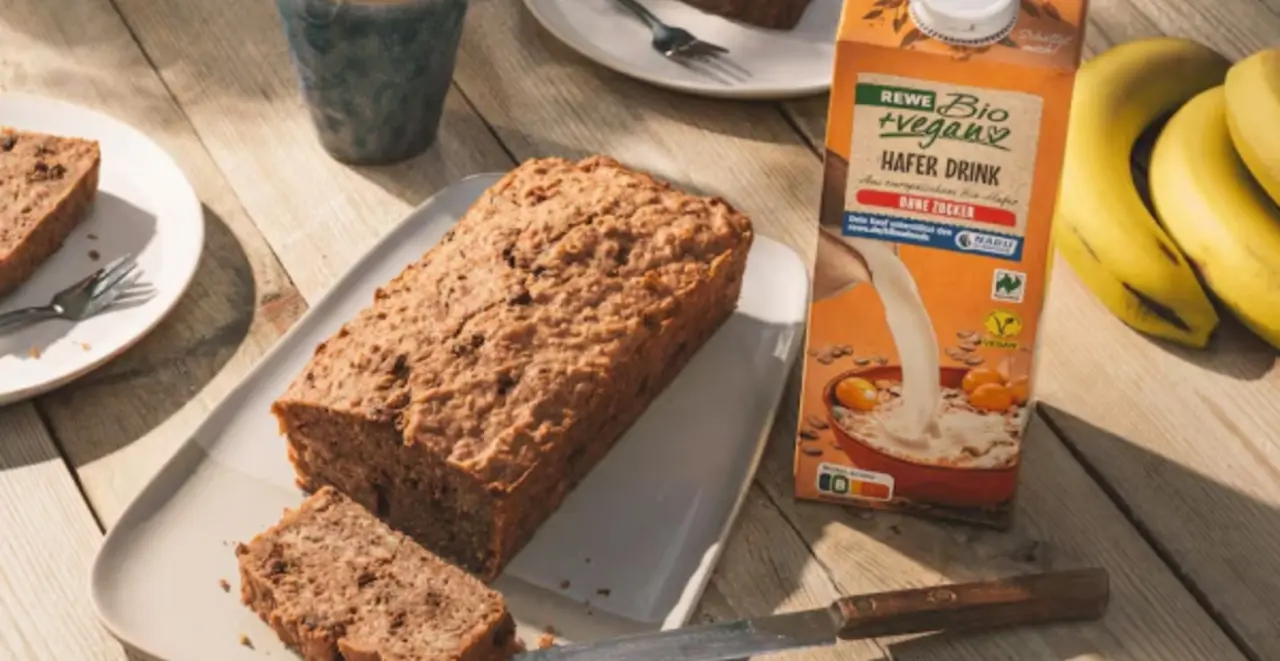 Ein angeschnittetes Bananenbrot auf einer Platte, zwei Teller, auf denen Brotscheiben liegen. Rechts davon eine Packung REWE Bio Hafer Drink.  