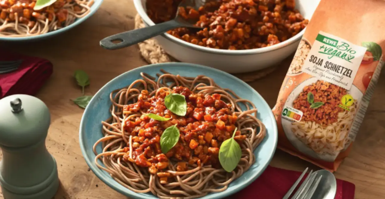 Ein Teller Vollkorn-Spaghetti mit veganer Bolognese und frischem Basilikum. Oberhalb steht eine Pfanne mit Bolognese, rechts eine Packung REWE Bio Soja Schnetzel.