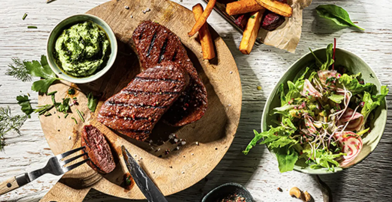 Auf einem Holzbrett stehen gegrillte pflanzliche Steaks mit einem kleinen Salat und Dipp.